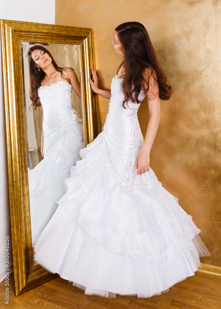Fototapeta premium Beautiful dark haired woman in white bridal dress standing nearby mirrow and looking at it