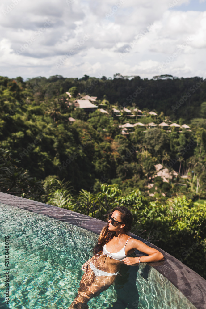 Woman in white swimsuit and sunglasses relaxing in luxury infinity pool with jungle view in Ubud, Bali. Luxury lifestyle, amazing vacations in Bali.