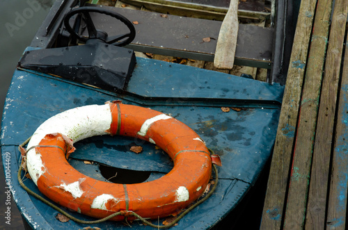 A lifeboat moored near a wooden pier. Inside is a wooden oar and a life preserver. An old motorboat with a steering wheel. Poor working conditions. Use of old equipment.