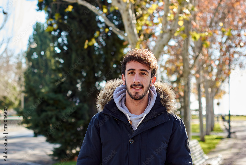 Portrait of young man with beard, looking at camera with background of winter trees and sun lighting between the leaves. Trees in winter with colorful leaves Portrait of young man with beard, looking at camera with background of winter trees and sun lighting between the leaves. Trees in winter with colorful leaves