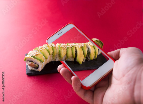 A hand holds a phone and photographs green sushi with avocados and this roll goes out of the scope of the phone. Close-up photo.