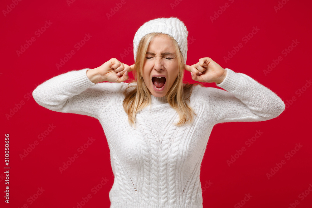 Frustrated young woman in white sweater, hat isolated on red background. Healthy fashion lifestyle cold season concept. Mock up copy space. Keeping eyes closed, screaming, covering ears with fingers. Frustrated young woman in white sweater, hat isolated on red background. Healthy fashion lifestyle cold season concept. Mock up copy space. Keeping eyes closed, screaming, covering ears with fingers.