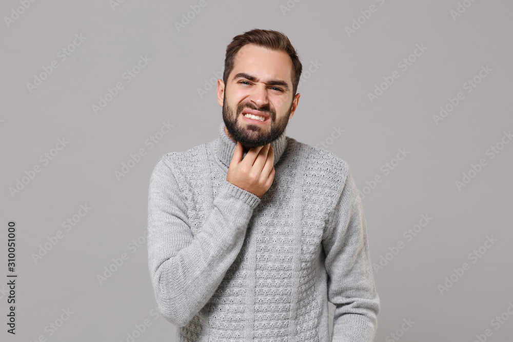 Young bearded man in gray sweater posing isolated on grey wall background, studio portrait. Healthy lifestyle, ill sick disease treatment, cold season concept. Mock up copy space. Put hand on throat. Young bearded man in gray sweater posing isolated on grey wall background, studio portrait. Healthy lifestyle, ill sick disease treatment, cold season concept. Mock up copy space. Put hand on throat.