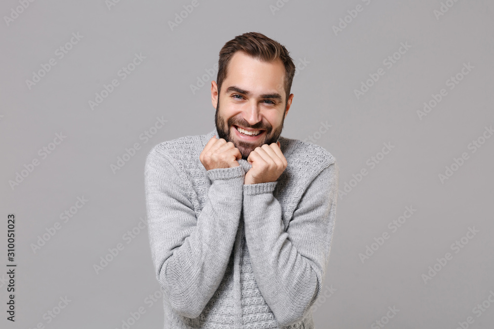Smiling frozen young bearded man in gray sweater posing isolated on grey background studio portrait. Healthy fashion lifestyle people emotions cold season concept. Mock up copy space. Clenching fists. Smiling frozen young bearded man in gray sweater posing isolated on grey background studio portrait. Healthy fashion lifestyle people emotions cold season concept. Mock up copy space. Clenching fists.