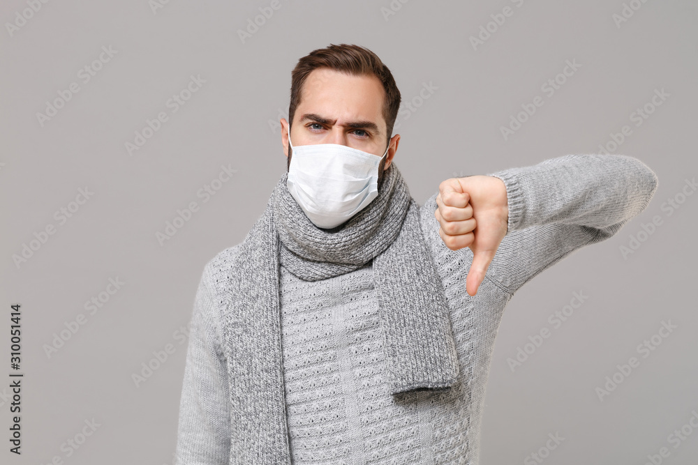 Young man in gray sweater, scarf isolated on grey background. Healthy lifestyle, ill sick disease treatment, cold season concept. Mock up copy space. Wearing sterile face mask, showing thumb down. Young man in gray sweater, scarf isolated on grey background. Healthy lifestyle, ill sick disease treatment, cold season concept. Mock up copy space. Wearing sterile face mask, showing thumb down.