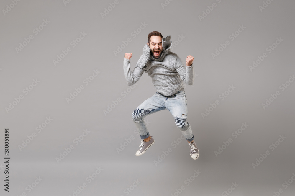 Happy young bearded man in gray sweater, scarf posing isolated on grey background, studio portrait. Healthy fashion lifestyle, cold season concept. Mock up copy space. Jumping, doing winner gesture.