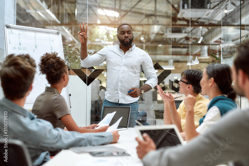 One team one goal. Confident afro american businessman standing near whiteboard and explaining something to his young colleagues while working together in the creative office