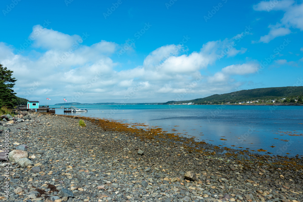 Rocky beach on a sunny day with blue sky and clouds. The water is teal blue and smooth. There's a mountain in the background.