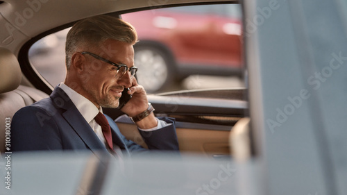 I am always available. Side view of cheerful mature man in classic wear sitting in the car and talking by the phone with client