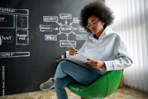 Reading at work. Young afro american female graphic designer in eyeglasses reading magazine while sitting on the chair against blackboard in the big creative office. Creative agency