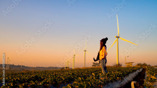 One woman stands in the middle of a wide field have large wind turbines, Which is an industry that produces electricity from clean energy.
