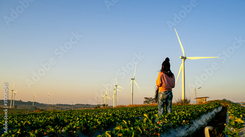 One woman stands in the middle of a wide field have large wind turbines, Which is an industry that produces electricity from clean energy.