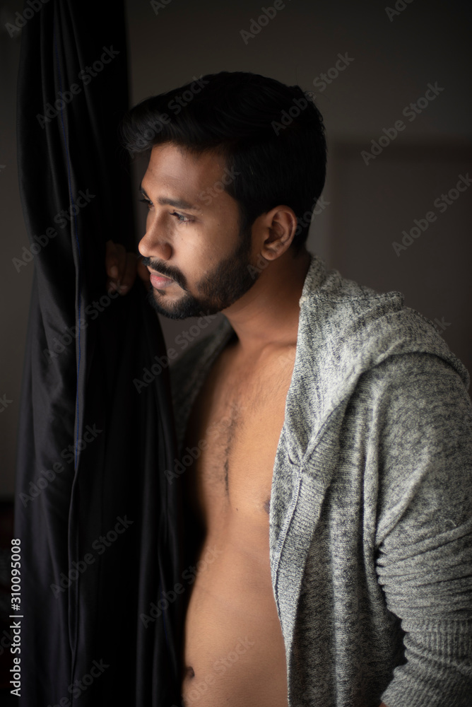 An young tall, dark and handsome Indian Bengali man in a front open western jacket looking outside white standing in front of a window in studio background. Indian lifestyle and fashion.