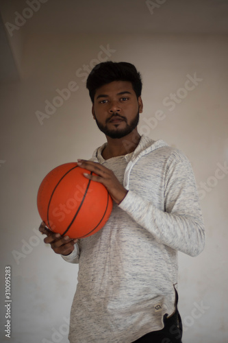 An young tall, dark and handsome Indian Bengali man in a western jacket and jeans with a basket ball in white background. Indian lifestyle and fashion.