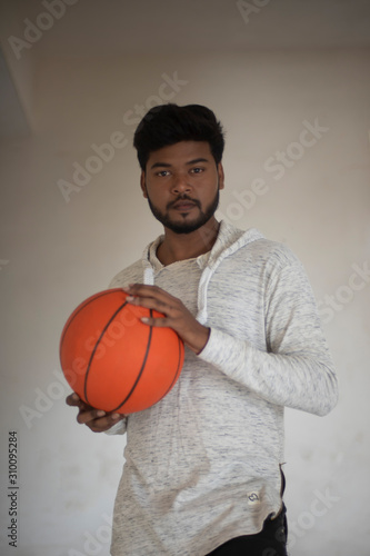 An young tall, dark and handsome Indian Bengali man in a western jacket and jeans with a basket ball in white background. Indian lifestyle and fashion.