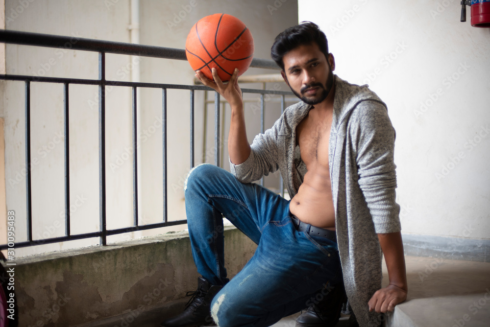 An young tall, dark and handsome Indian Bengali man in a front open western  jacket and jeans with basket ball in white background. Indian lifestyle and  fashion. Stock Photo