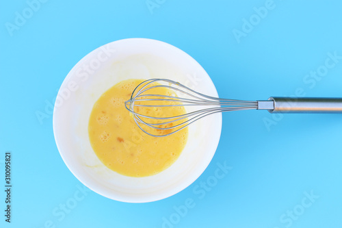 Broken eggs in a bowl and an iron whisk on blue background close-up.