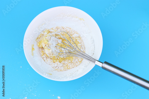 flour with an egg in white bowl and an iron whisk top view of blue background.