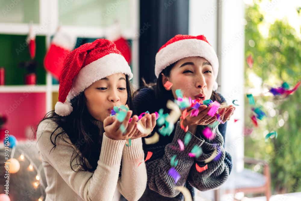 Cute girl and mother in santa hats having fun blowing glitter and smiling while celebrating new year eve and enjoying spending time together in christmas time at home Cute girl and mother in santa hats having fun blowing glitter and smiling while celebrating new year eve and enjoying spending time together in christmas time at home