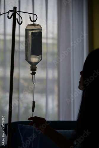 Silhouette picture of patient is looking at saline fluid bag and holding saline tube with white curtain background.