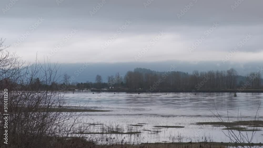 Blue river flowing on a cloudy day with bare branches and dry grass in the foreground and mountains in the background (normal speed) Blue river flowing on a cloudy day with bare branches and dry grass in the foreground and mountains in the background (normal speed)