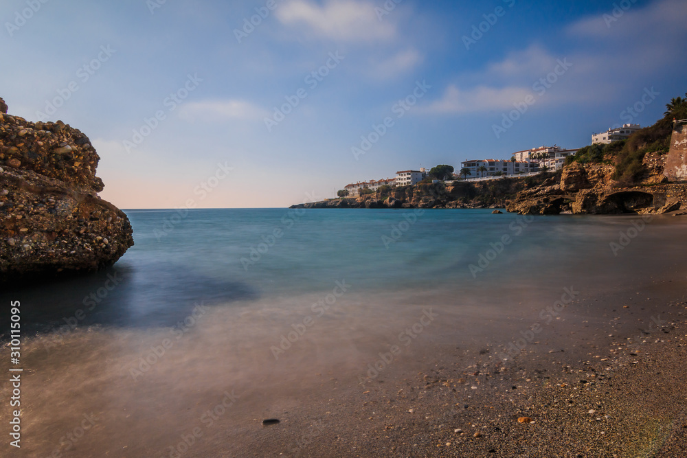 Beach section on the Mediterranean coast in Nerja. Sandy beach on the Spanish coastline of Costa del Sol with rocks on sunny day with blue sky and clouds