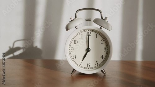 White alarm clock is placed on a wooden table and the light shines through the curtains in the morning.