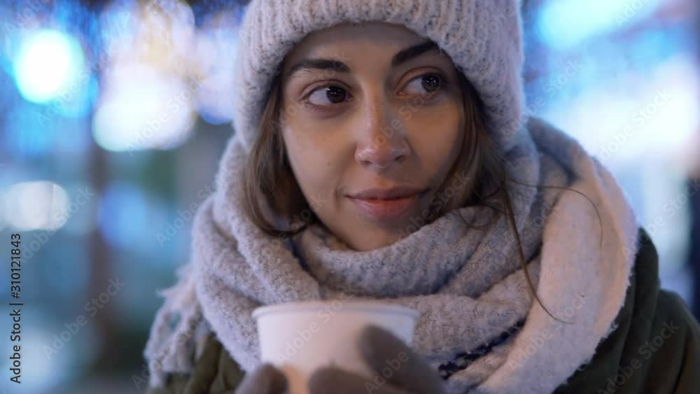 attractive young woman in white woolen cap and scarf standing outdoors on illuminated winter city street at evening and drinking coffee from paper cup. woman takes a sip and enjoys the aroma and taste attractive young woman in white woolen cap and scarf standing outdoors on illuminated winter city street at evening and drinking coffee from paper cup. woman takes a sip and enjoys the aroma and taste