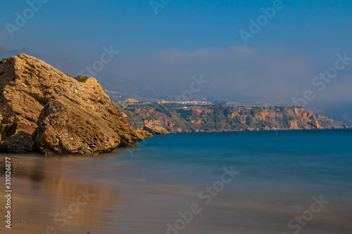 Coastal section on the Spanish Mediterranean coast in Andalusia. Costa del Sol with rocks and blue water in sunshine. Blue sky and cityscape in the background
