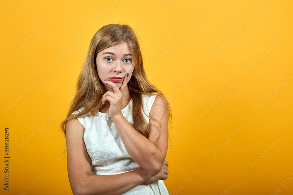 Serious caucasian blonde young woman keeping finger on cheek, looking dissatisfaction over isolated orange background wearing white shirt. Lifestyle concept Serious caucasian blonde young woman keeping finger on cheek, looking dissatisfaction over isolated orange background wearing white shirt. Lifestyle concept