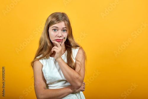 Serious caucasian blonde young woman keeping finger on cheek, looking dissatisfaction over isolated orange background wearing white shirt. Lifestyle concept