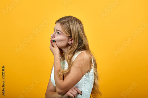 Disappointed blonde young caucasian woman holding finger on cheeks, keeps lips clamped over isolated orange background wearing white shirt. Lifestyle concept