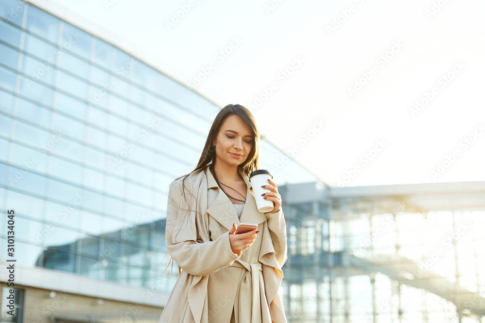 Beautiful smiling woman walking on near office from work with coffee cup and texting on mobile phone.Phone communication. Beautiful smiling woman walking on near office from work with coffee cup and texting on mobile phone.Phone communication.