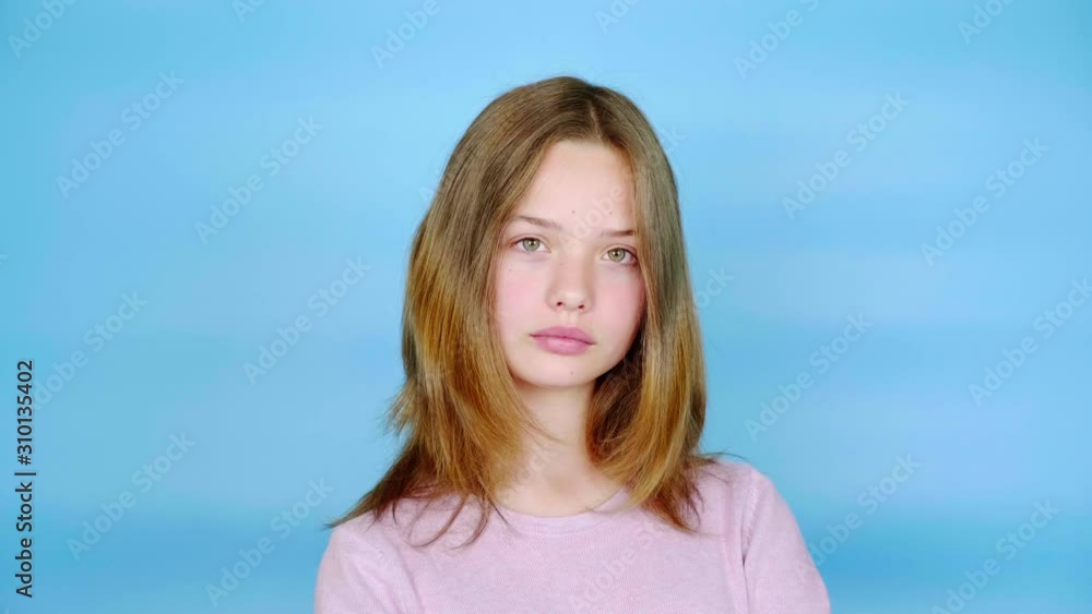 Teen girl in a pink pullover is looking at the camera. Blue background with copy space. Teenager emotions. 4k footage Teen girl in a pink pullover is looking at the camera. Blue background with copy space. Teenager emotions. 4k footage