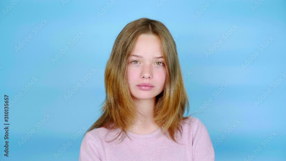 Teen girl in a pink pullover is looking at the camera. Blue background with copy space. Teenager emotions. 4k footage Teen girl in a pink pullover is looking at the camera. Blue background with copy space. Teenager emotions. 4k footage