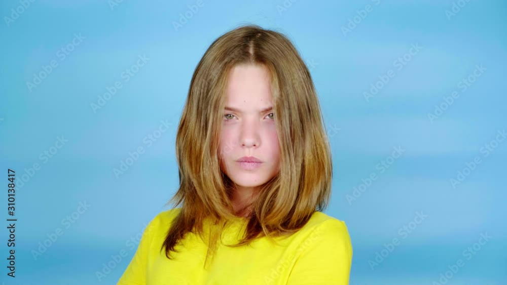 Teen girl in a yellow t-shirt is angry and looking at the camera. Blue background with copy space. Teenager emotions. 4k footage Teen girl in a yellow t-shirt is angry and looking at the camera. Blue background with copy space. Teenager emotions. 4k footage