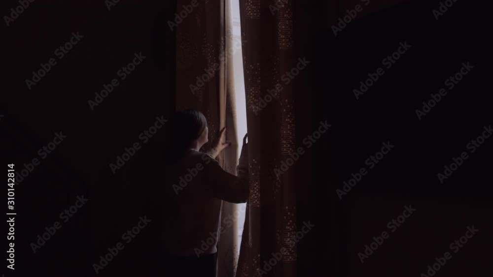 Girl opens curtains looks out window in dark room silhouette of young woman sunlight Girl opens curtains looks out window in dark room silhouette of young woman sunlight