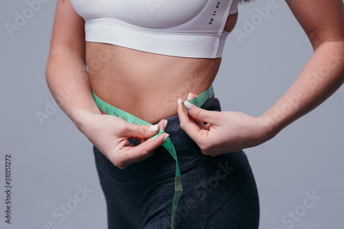 Slender young woman measures her slim waist with a tape measure, closeup in a white tank top and gray leggings