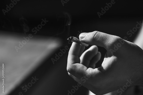 A black-and-white image of a hand holding a cigarette butt emitting smoke and illuminated by a bright backlight.