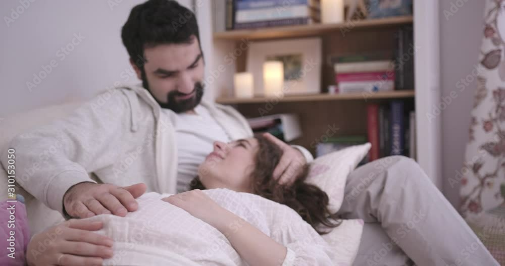Man and Pregnant Woman Sitting lying on sofa, Embracing and Laughing. Young Family Expecting a Baby. White Interior with Baby Cot on the background. Concept of Parenthood, Happiness and Tenderness Man and Pregnant Woman Sitting lying on sofa, Embracing and Laughing. Young Family Expecting a Baby. White Interior with Baby Cot on the background. Concept of Parenthood, Happiness and Tenderness