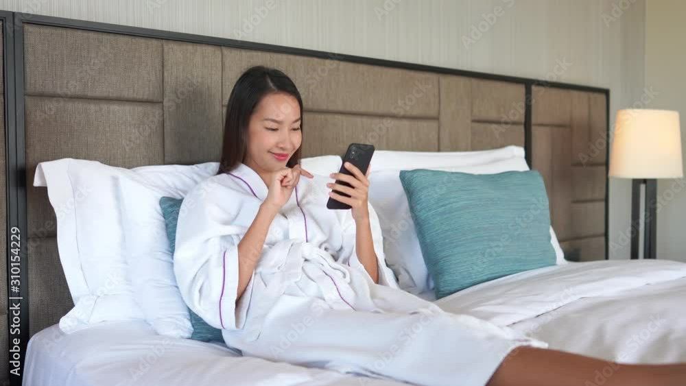 Delighted asian lady scrolling on her phone on bed in a bathrobe