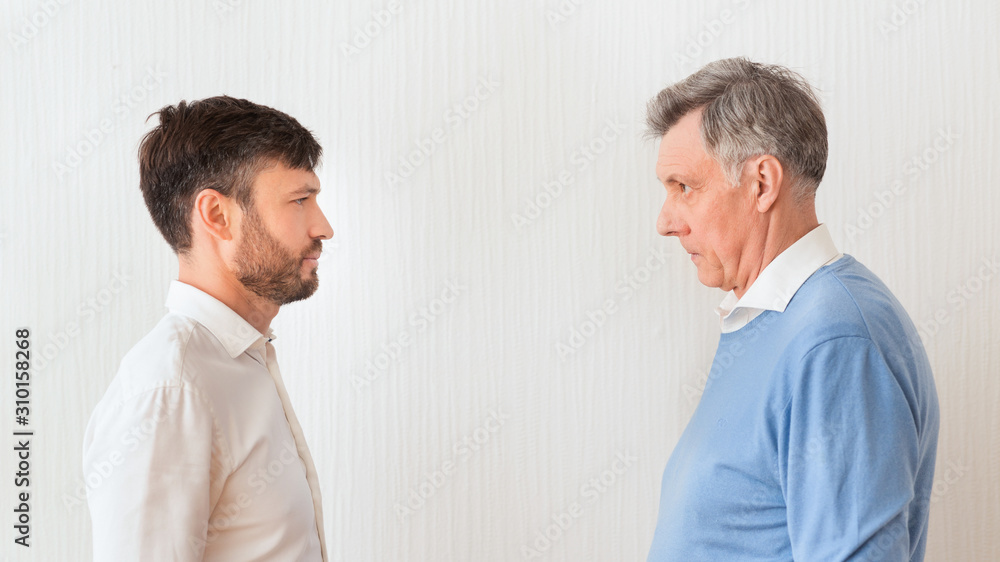 Elderly Man And Son Looking Severely Standing Face-To-Face Against Wall Elderly Man And Son Looking Severely Standing Face-To-Face Against Wall