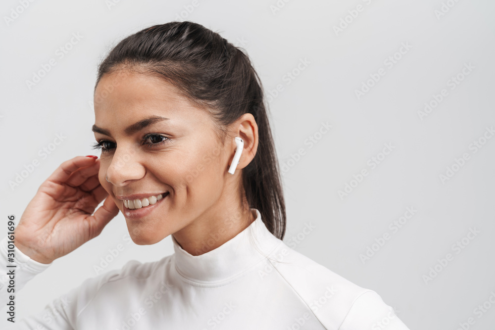 Image of young brunette woman in sportswear putting on earpods isolated over gray background Image of young brunette woman in sportswear putting on earpods isolated over gray background