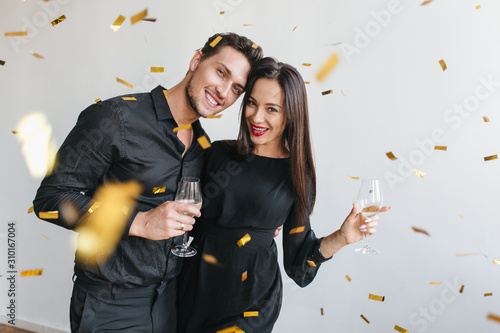 Handsome brunette man embracing his girlfriend and looking at golden confetti at party. Dark-haired lady in elegant dress celebrating something with husband and drinking champagne.