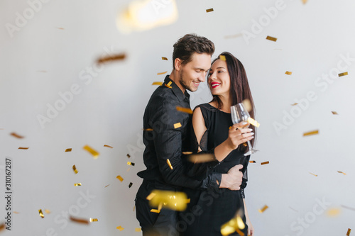 Brunette long-haired woman looks at husband's face with tenderness, enjoying birthday party. Indoor portrait of loving couple celebrating anniversary with sparkle confetti on foreground.