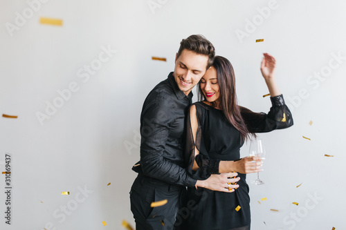 Stylish loving couple dancing together at party, looking at sparkle confetti falling down. Charming young woman in black attire fooling around with boyfriend during event.