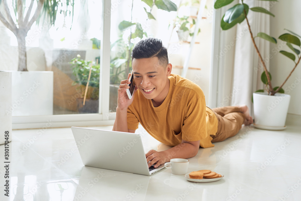 Smiling man talking on cellphone and using laptop while lying on a floor at home Smiling man talking on cellphone and using laptop while lying on a floor at home