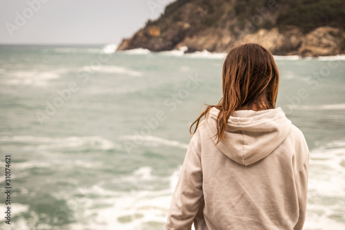 Young woman with sweatshirt sitting in front of the sea looking towards the horizon. There is a mountain in the background while breaking a wave in front of the woman.
