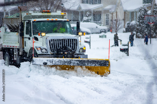 Front view of city services snow plow truck with yellow blade cleaning roads after winter storm with kids playing in the background