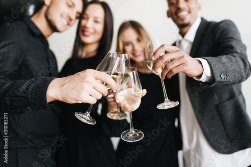Smiling european man leaning to black-haired wife during party photoshoot. Blur portrait of african guy posing with friends at home fest with wineglasses on foreground.